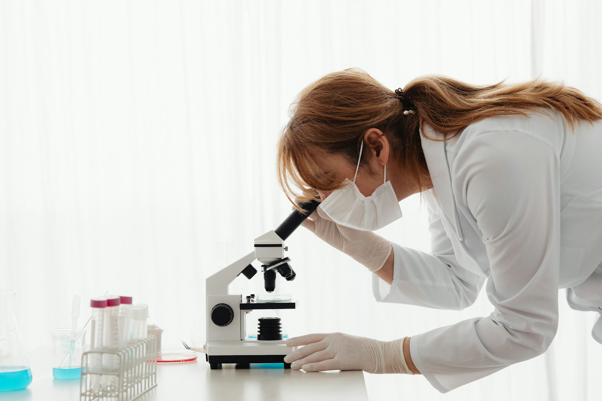 Biotehnolog, stručnjakinja za biotehnologiju s maskom na licu, u bijeloj kuti, u laboratoriju gleda u mikroskop, epruvete su pored