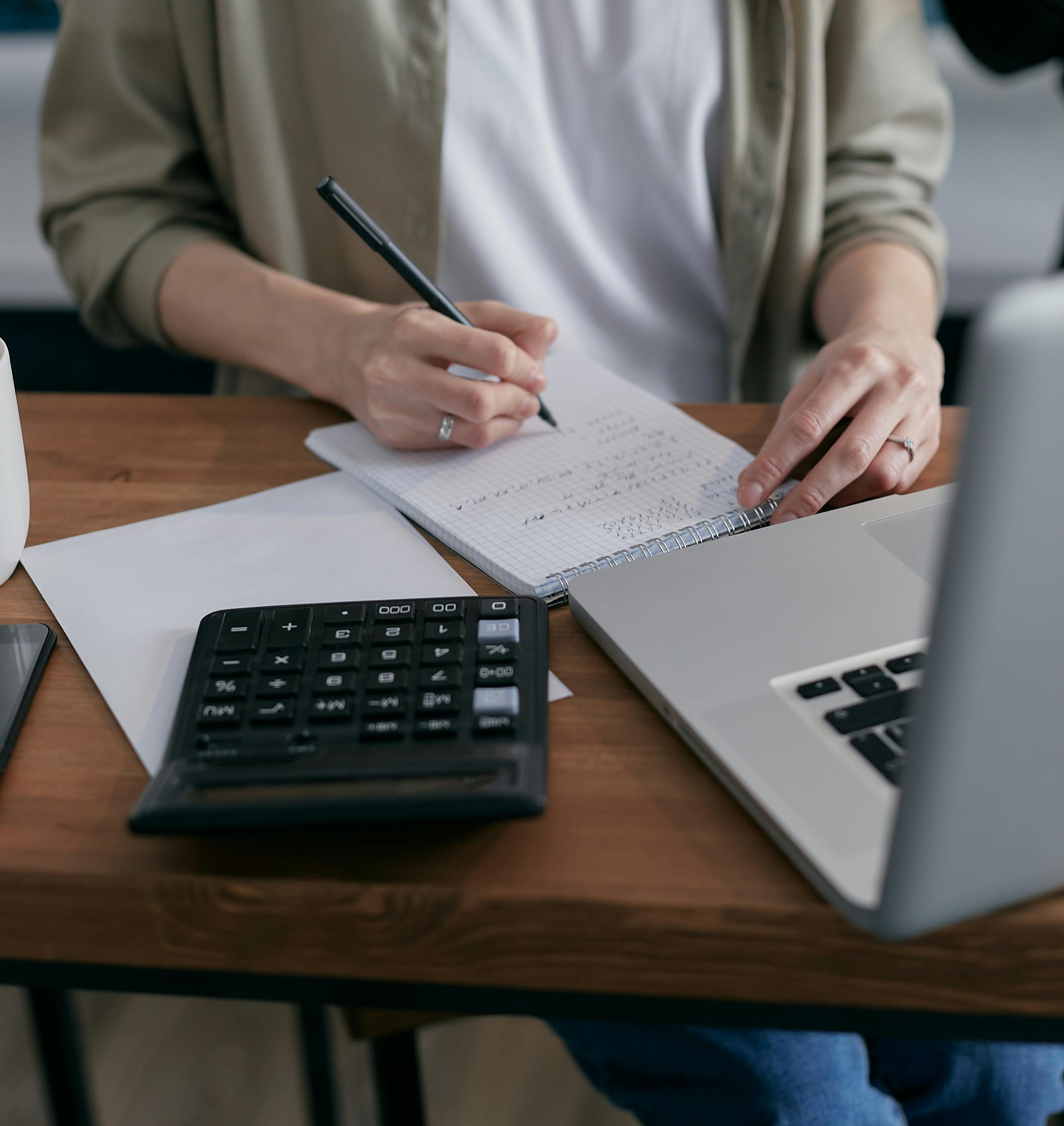 Financijski analitičar sjedi za radnim stolom, piše po bloku, ispred su kalkulator i laptop