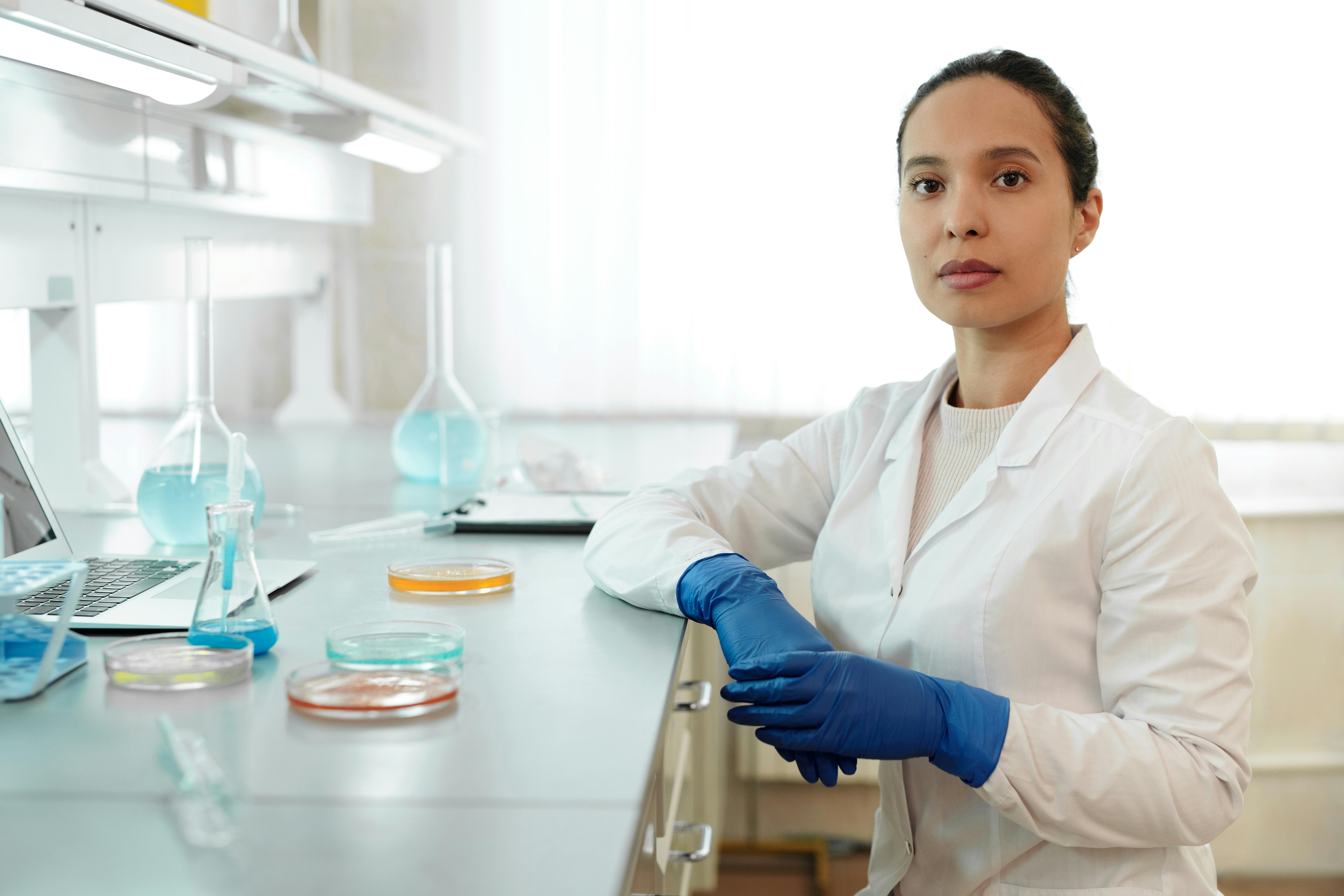 Klinički istraživač, istraživačica, sjedi u laboratoriju, ima bijelu kutu i plave rukavice, ispred nje su epruvete